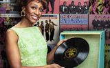 Beverley Knight stands in front of a wall of album cover artwork for The Drifters. She's holding a record about to play it on a vintage record player