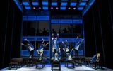 Cast members perform in school uniform amongst a school classroom set. Cast stand on chairs and desks