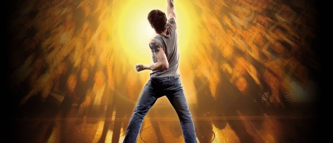 Man wearing jeans stands looking out from the stage facing out to the audience with one arm punching the air