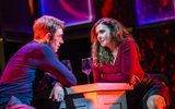 A man and a woman talk at a table with red lighting