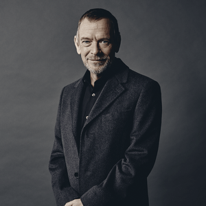 A man wearing a grey suit jacket and dark shirt stands and looks at the camera