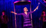 A glamorous woman in a purple dress takes the applause from the audience whilst holding a magic wand with a star at its tip