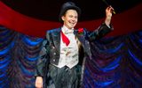 A smiling magician wearing a top hat and tails waves to the audience