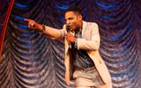 A man in a smart light coloured suit points to the audience whilst talking into a microphone on stage