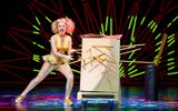 A glamorous assistant in a gold costume has an exaggerated expression on her face as she plunges long stakes through a wooden box