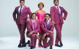 A woman in a glitzy bright pink dress is flanked on either side by two members of a male vocal group dressed in matching purple suits with two further members kneeling before her wearing similar coloured outfits