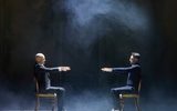 Two men sitting on chairs opposite each other stretch out their arms