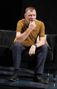 A man in a mustard coloured t-shirt sits holding his chin in his hands looking worried