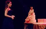 A woman in a glamorous dark blue dress stands in front of a large birthday cake