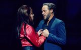 A woman with long dark hair passionately grasps the hand of a handsome man in a smart blue suit