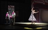 A woman in a floral dress sings on stage while poeple wearing Hawaiian leis stand beside her.