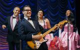 A male and two female backing singers stand with a man in a smart, dark coloured suit holding a guitar