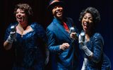 Two female singers in glamorous blue dresses stand either side of a man in a dark blue suit and hat as they perform