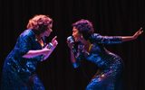 Two female singers in sparkly blue dresses face either other as they perform a song