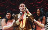 A man wearing an animal print jacket holds a telephone to his ear as he is flanked by two female performers