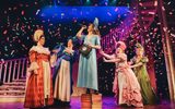 A woman stands on a large pile of books whilst singing into a microphone whilst four other women look on with amusement whilst being showered with ticker tape