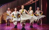 Five women perform a song and dance routine whilst wearing white petticoats and yellow washing up gloves