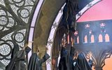 A nun swings from a long rope in front of a stained glass window with a look of horror on her face. A group of nuns stand below and hold their arms up in the air towards her.