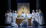 A group of nuns dressed in white nightgowns surround a bed and listen intently to a woman sitting on the bed