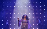 A woman stands centre stage under a spotlight and performs a song dressed in a glamorous sparkly dress