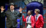 A boy and girl wearing red school cardigans try to get the attention of a policeman who is standing underneath a street lamp