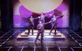 Four men in purple suits holding microphones with their arms outstretched as they are dancing.