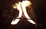 One male stands on stage with his arms in motion with huge streaks of fire above him.