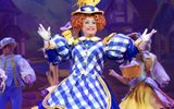 The pantomime dame smiles looking out into the audience, they are wearing a blue and white checked dress and hat.