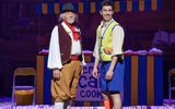 Two males standing opposite each other, the elder of the two wears a hat and is dressed in a red waistcoat and brown trousers. The younger of the two wears a bright green waistcoat and blue shorts. They are both smiling cheekily,