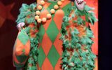 The pantomime dame wears an orange and green patterned dress with a large orange necklace and feathers on the sleeves.