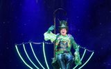 Joe McElderry dressed in a green sparkly outfit sits on an illuminated umbrella as it hovers mid air.