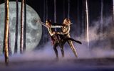 Two dancers - one blindfolded - are surrounded by tall trees in a mysterious forest