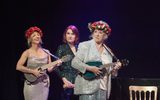 Three women all in sequined outfits, two wearing flower hairbands and carrying ukuleles.