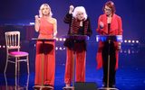 Three females in red and black outfits standing in front of music stands with their hands making fists.