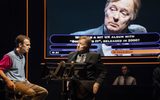 A man in a suit sits opposite a male in jeans and a t-shirt. The face of the male wearing the suit is projected onto a question screen behind the pair.