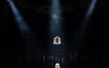 A man in smart dress stands under spotlights in an otherwise pitch black room.
