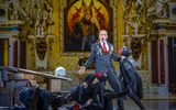 A male wearing a suit sings whlst others surround him wearing masks.
