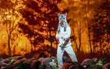 A male dressed as a wolf stands in front of a flamed background whilst lots of bodies are on the floor surrounding him.
