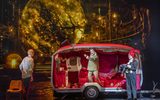 Three males look accusingly at each other standing in front of a caravan decorated in red velvet.