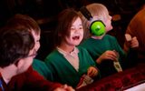 A girl is laughing while holding a packet of crisps, sat beside someone wearing ear defenders.