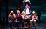 Four people sit on chairs wearing rainhats while a man stands over them holding a fish above his head.