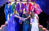 4 women wearing colourful dresses hold their hands together while 5 young men wearing colourful suits stand over them looking excited