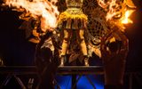 A woman stands in a black and gold dress looking serious with her arms raised above her head. Pyrotechnics go off beside her.