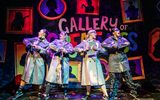 Four females wearing long trench coats stand in front of a sign reading 'Gallery of Greatness'.