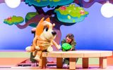 A cartoon dog sits on a wooden outdoor table next to a leafy green tree. The dog is talking to a small green insect.