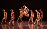 One male jumps in the air with his feet touching his head, another six dancers stand behind him looking up.