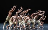 A large group of dancers all facing the side and leaning slightly forwards with their arms outstretched.