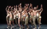 A large group of dancers all facing the front and leaning slightly backwards with their arms above their heads whilst balancing on one leg.