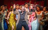 A male in a flamboyant leopard print suit sings and dances whilst surrounded by people in 1905s style dresses.