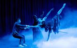 A female balances on one leg whilst she kicks the other behind her. The bottom of the stage is filled with smoke and it is dimly lit.